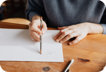 A flat wooden surface with a sheet of white paper on it, with a person, visible from chest down, behind it. The person's right hand is holding a pencil to the paper, where faint markings can be seen.